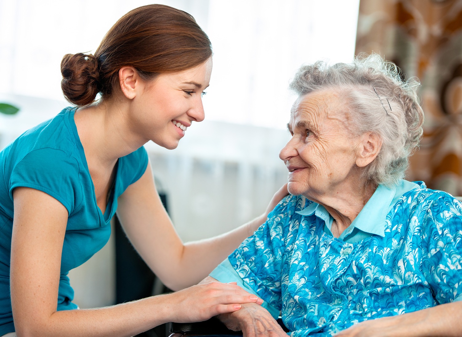 Young woman providing in-home care to elderly woman, both looking at each other smiling while elderly woman is seated