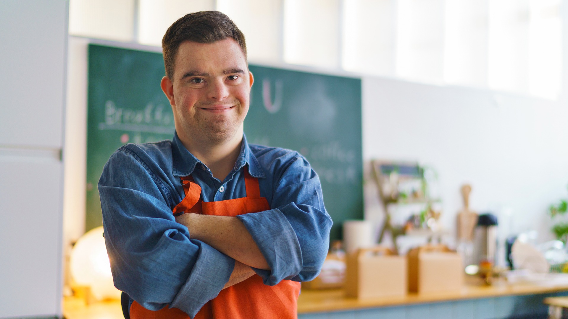 A young man with Down syndrome working in sandwich shop smiling at the camera with arms crossed