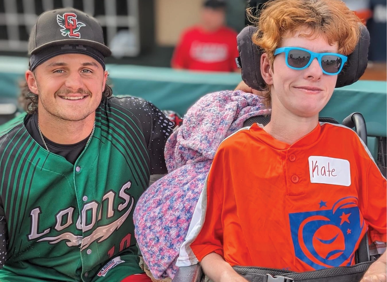 Great Lakes Loons Baseball Player kneeling next to woman using wheelchair, both are smiling at the camera for a photo.